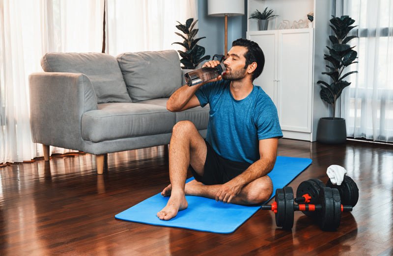 Man in blue shirt drinking water on blue exercise mat at home with dumbbells and couch nearby